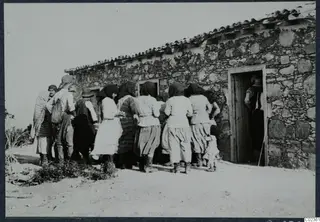 Women and men gathered outside a stone building with a wooden door at Vouni, waiting for payment.