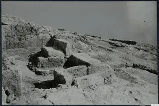 Excavated stone walls and blocks on a rocky slope at Vouni, with a dark cut opening in the foreground.