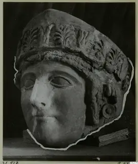 Sculpted head of a figure wearing a tall headdress carved with leaf motifs, shown on wooden blocks against a dark backdrop.