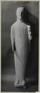 Limestone statue of a standing figure in a long garment, one hand holding an object at the chest, head and feet partly worn.