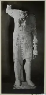 Headless limestone statue of a standing figure in a short pleated garment, on a base
