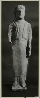 Limestone statue of a standing figure in a draped cloak, arms at its sides, shown against a dark backdrop