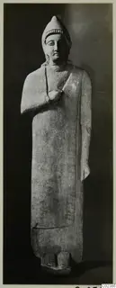 Limestone statue from Kition of a standing figure in a tall cap and long robe, one hand raised to the chest.
