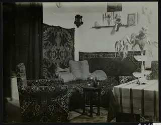 Interior of a saloon with a patterned sofa, cushions, a wall shelf with a doll and photographs, and a draped table