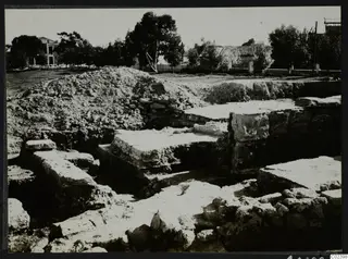 Excavated stone walls and large cut blocks at Bamboula, Kition, with a spoil heap behind and trees beyond.