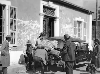 Men and a woman loading striped bedding, a basket and suitcases onto an old open car outside a building in Nicosia