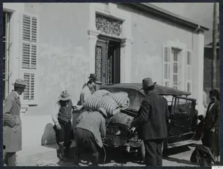 Men loading bundles, baskets and suitcases onto an old car outside a building in Nicosia