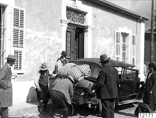 Men loading bundles and luggage onto an old car outside a plastered building with shuttered windows, Nicosia