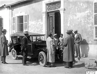 Men in long coats and hats stand beside a dark open car outside a plastered building in Nicosia