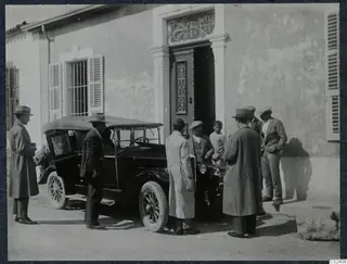 Men in coats and hats stand beside an open-top car outside a building with an ornate iron grille over its doorway.