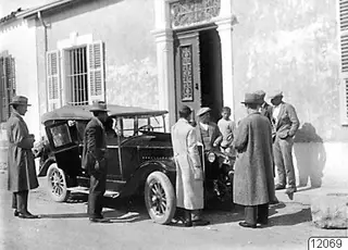 Men in long coats and hats standing beside an open-topped car outside a plastered building in Nicosia