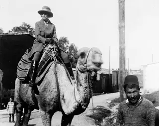 Vivi Gjerstad riding a camel on a dirt road in Nicosia, with a man leading the camel beside her.