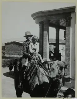 Vivi and Stefan Gjerstad riding a camel past a stone-columned porch