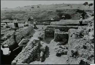 Excavation at the L.C. III house, Ayia Irini, with workers among exposed stone walls and a wheelbarrow of earth.