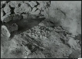 In situ finds on an excavated floor at Ayia Irini: a cluster of small stones and broken vessel pieces beside a rough stone wall.