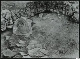 Excavation at Ayia Irini showing a curved stone wall, a large partly buried vessel and scattered sherds.