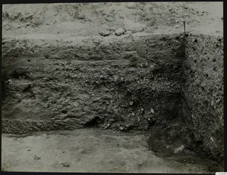 Excavated trench section at Ayia Irini showing layers of soil and stones, with a wooden stake at the top edge.