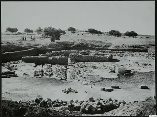 Excavation at Ayia Irini with low stone walls and a squared stone block, with people and trees in the background.