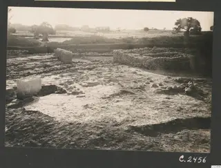 Excavated stone walls and a paved floor at Ayia Irini, with a stone block at left and trees and buildings beyond.