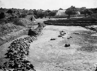 Excavated area at Ayia Irini with a long stone wall on the left, a tent and a white building behind.