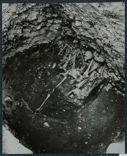 Skeleton lying in an excavated pit at Enkomi, with a skull and long leg bones visible and stones scattered on the floor