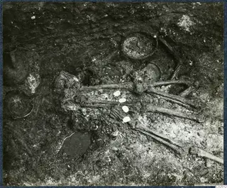 Tomb chamber at Enkomi during excavation, with a skeleton lying in dark soil among pottery vessels