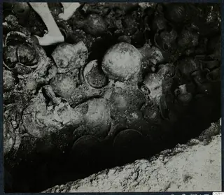 Tomb 13 at Enkomi: a dense cluster of pottery vessels in the tomb chamber during excavation, with a person's legs at the top edge.