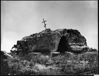 Built stone tomb with a dark doorway opening, a wooden cross on top, set in grass and scrub