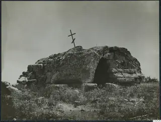 Built tomb of large stone blocks with a dark entrance opening, a wooden cross on top, grass in the foreground