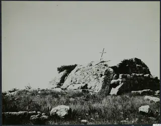 Built stone structure with a wooden cross on top, set among grass and boulders