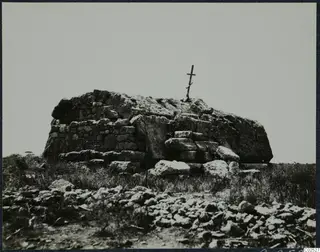 Built stone tomb with a wooden cross on top, surrounded by grass and scattered stones.