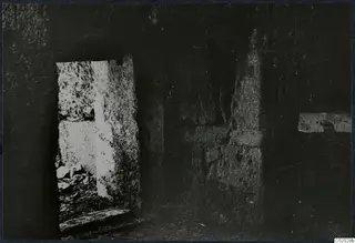 Interior of a built tomb chamber during excavation, with stone walls, a bright doorway on the left and rubble on the floor