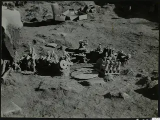 Terracotta figurines and carts lying in excavated ground at Ayia Irini, with cardboard boxes behind.