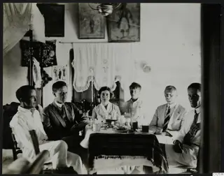 Six people seated around a dining table in a whitewashed room with framed paintings and a lace curtain on the wall.