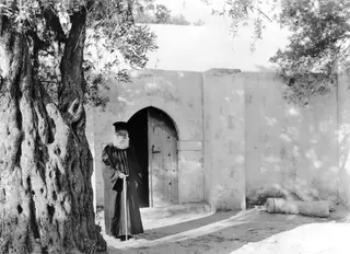 Papa Prokopios, a bearded man in a long dark robe and black hat, standing by an arched doorway at Ayia Irini