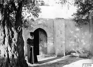 Papa Prokopios standing with a walking stick beside an arched doorway in a whitewashed wall at Ayia Irini