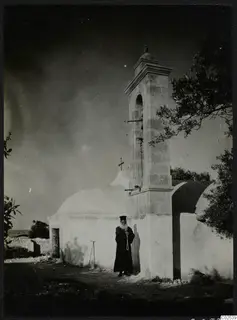 Papa Prokopios in black robes ringing the bell of the whitewashed chapel at Ayia Irini.
