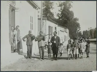 Two men play violin and lute in a street outside a whitewashed building, with children gathered around and a man holding a cup.