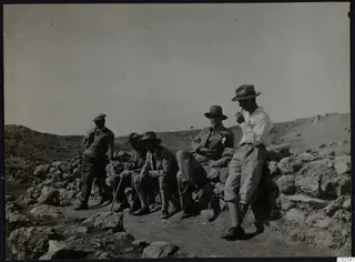 Five men in hats and jackets resting on and beside a rough stone wall in rocky hill country at Nitovikla.