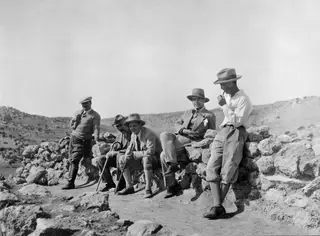 Five men sitting and standing on a rough stone wall in a dry, stony landscape, two of them holding walking sticks.