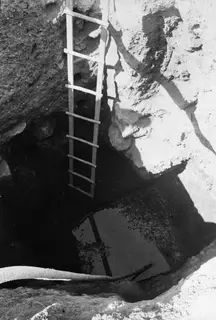 Tomb 18 at Enkomi seen from above: a wooden ladder leans down into the dark shaft beside a rock face.