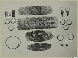 Gold and silver objects from Enkomi: thin decorated strips, rings, earrings and small cylinder seals laid out on a pale background.