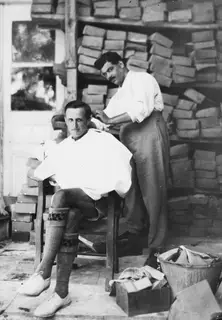 A seated man has his hair cut by a standing man, with stacked wooden boxes behind them, in Nicosia.