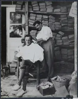 Man in a white cape having his hair cut indoors, in front of stacked wooden boxes.
