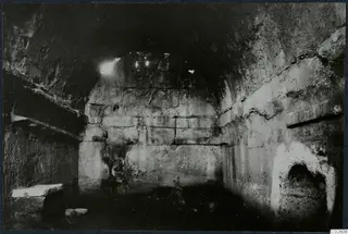 Interior of a built tomb chamber with a stone bench on the left and loose stones on the floor