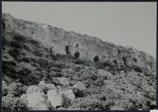 Pale limestone cliff with cut openings above a slope of bushes and boulders, Karpas peninsula