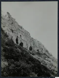 Steep pale limestone cliff with small arched openings cut into its face, with bushes on the slope below