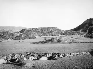 Necropolis hill at Palaeoskoutella seen from the south, with scrub-covered slopes rising behind open grassy ground.
