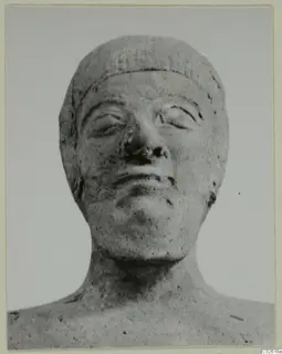 Frontal view of a carved head and upper shoulders with a broad band across the brow, against a plain background.