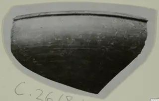 Sherd of a dark-surfaced vessel with a rounded body and a rim, photographed on a grey card mount
